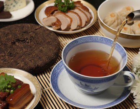 Traditional Chinese meal with tea, roasted duck, dumplings, pork belly, cheese, and chocolate on a bamboo mat.